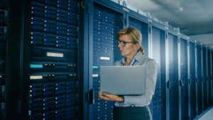 In Data Center: Female IT Technician Running Maintenance Programme on a Laptop, Controls Operational Server Rack Optimal Functioning. Modern High-Tech Telecommunications Operational Data Center.